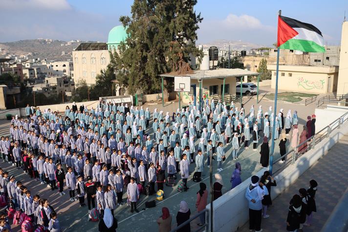 Hundreds of Palestinian students in uniform gathered for a morning assembly at a school courtyard with the Palestinian flag raised high.